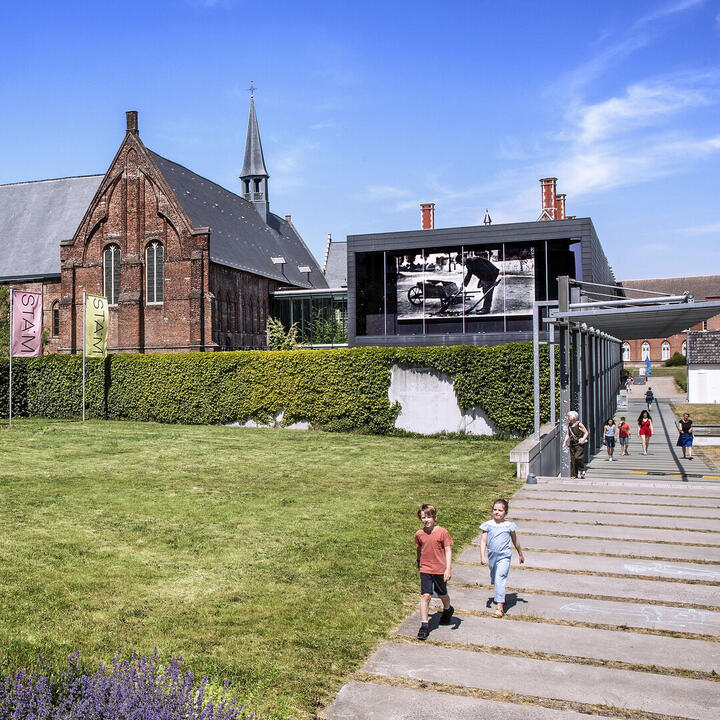 De voorgevel van het STAM, met een grasperk, een trap, wandelpad en vlaggen van het STAM.
