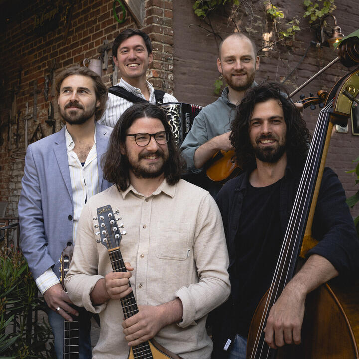 De afbeelding toont vijf muzikanten met verschillende instrumenten, waaronder een mandoline, een accordeon, een viool en een contrabas, staand voor een bakstenen muur met wat groen en een lantaarn