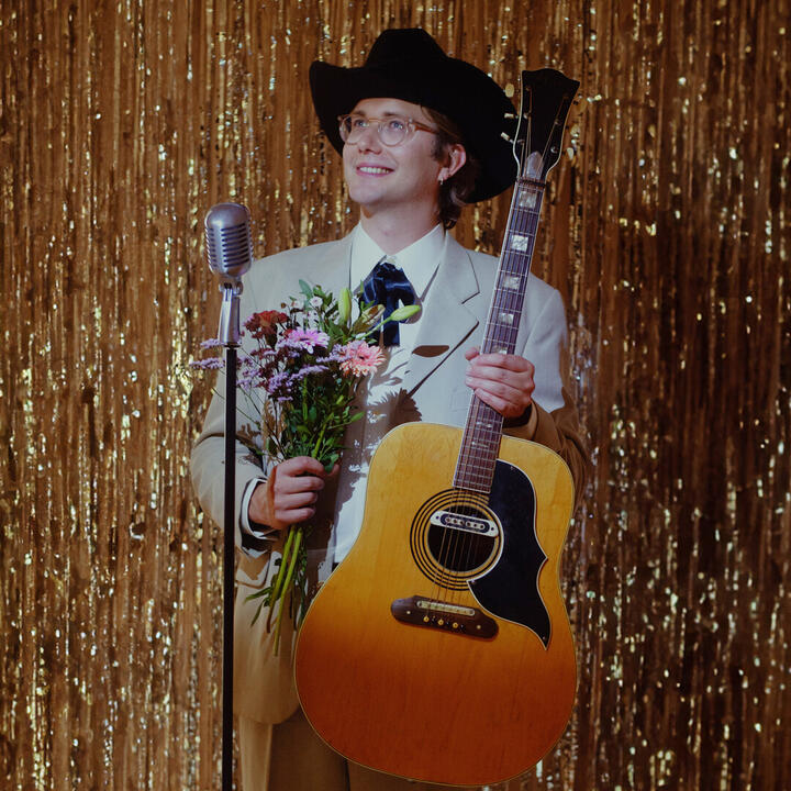 Een persoon in een lichtkleurig pak en een zwarte cowboyhoed, staand voor een gouden glitterachtergrond. Ze houden een akoestische gitaar en een boeket bloemen vast, met een vintage microfoon ervoor.