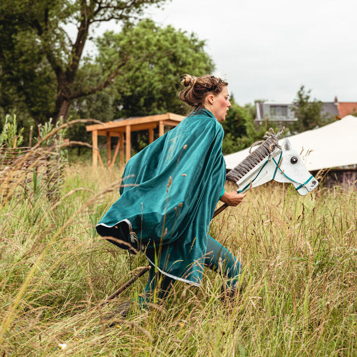 Een persoon in een turquoise mantel rent door hoog gras met een stokpaard onder de arm. Op de achtergrond staan bomen, een houten constructie en tentdoeken in een open veld.