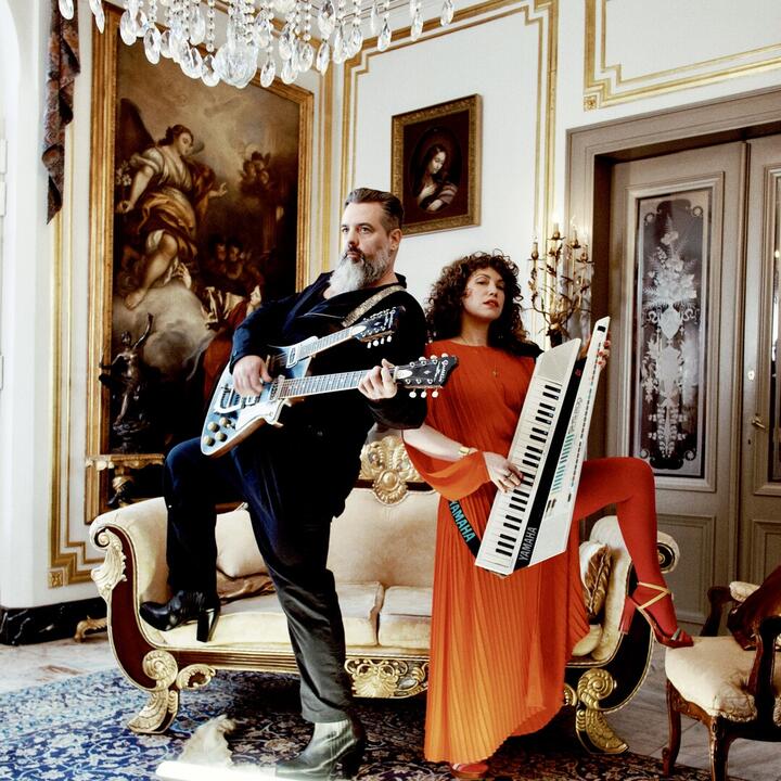 Twee personen in een rijkelijk versierde kamer, één met een gitaar en de ander met een Yamaha-keyboard.