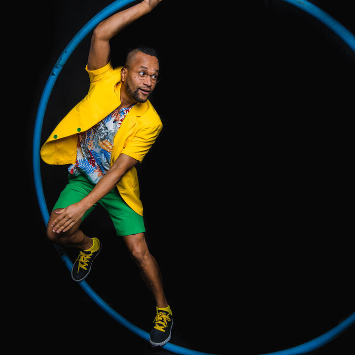 Een persoon voert een acrobatische pose uit met een grote blauwe hoepel, gekleed in felgekleurde kleding tegen een zwarte achtergrond.