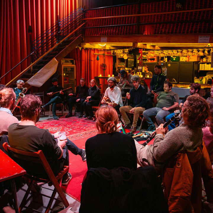 De afbeelding toont een groep mensen die in een cirkel zitten in een gezellige ruimte met warme verlichting, rode gordijnen en een trap naar een hoger niveau. De sfeer is intiem en sociaal.