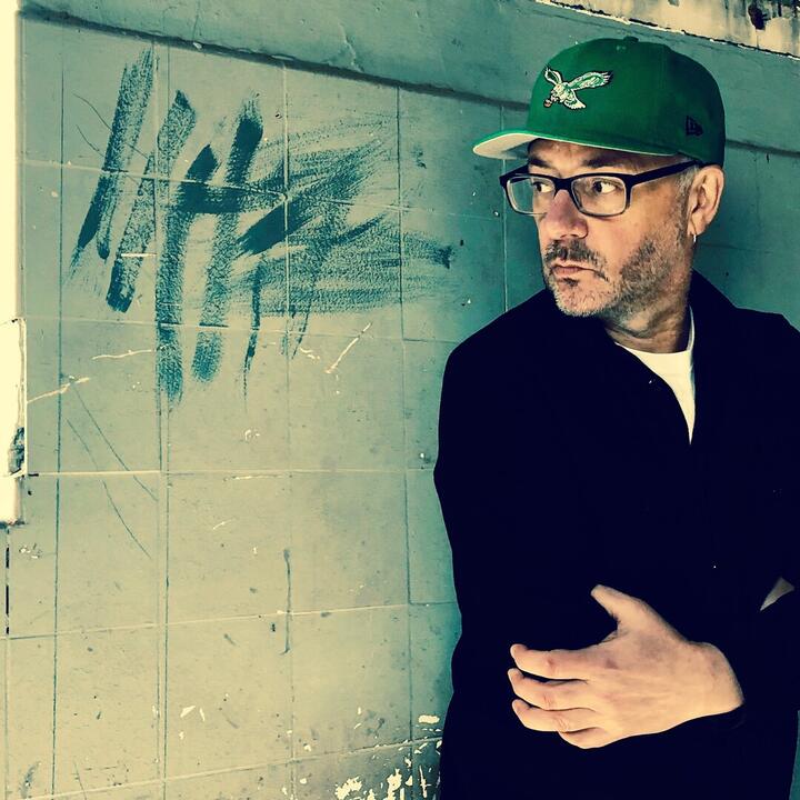 Een persoon staat tegen een betegelde muur met graffiti. Ze dragen een groene pet met een adelaar erop en een zwarte jas. Links is een raam met een rood object op de vensterbank. De omgeving lijkt oud en deels beschadigd