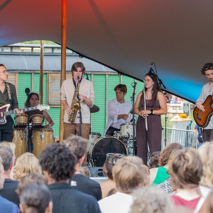 De band Ikaro staat op een podium. 2 mannen met een gitaar, een vrouw met een dwarsfluit, een mannelijke en vrouwelijke drummer en een man met een saxofoon.