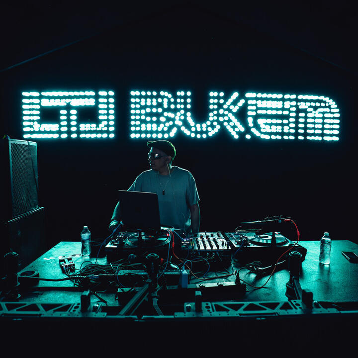 Een DJ treedt op achter een tafel met apparatuur, terwijl een verlicht bord met "LTJ BUKEM" op de achtergrond opvalt.