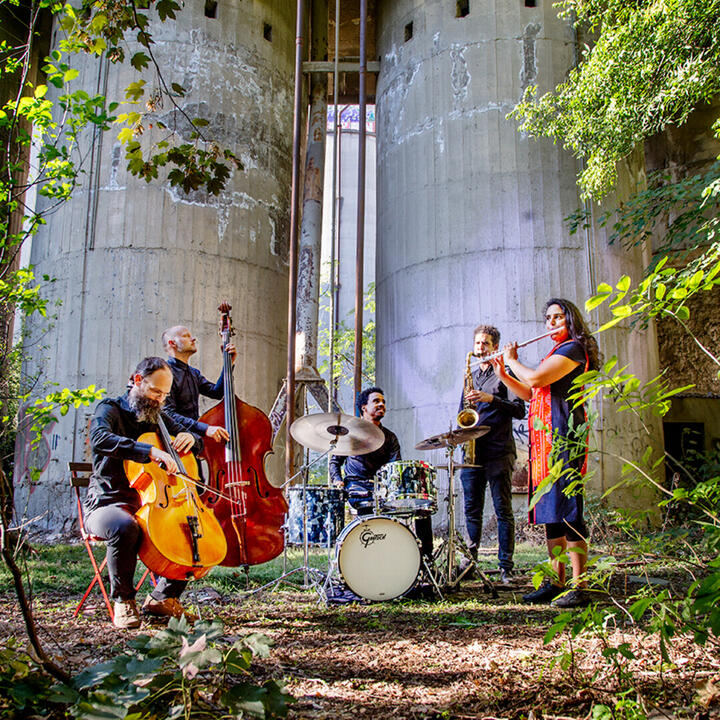 Vijf muzikanten spelen buiten in een bosrijke omgeving voor grote betonnen structuren. Ze bespelen een cello, contrabas, drumstel, saxofoon en fluit. De mix van natuur en industrie maakt de setting bijzonder.