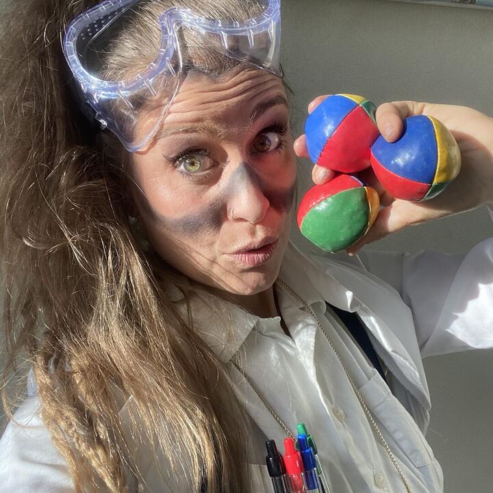 Een persoon draagt een veiligheidsbril en een witte overhemd met pennen in de borstzak, terwijl ze drie gekleurde jongleerballen vasthouden.