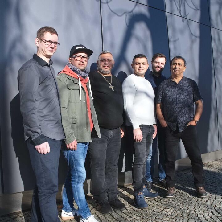 Zes mensen staan buiten op een stenen ondergrond voor een modern gebouw met een blauwgrijze muur. Hun schaduwen en die van boomtakken vallen op de muur. Ze dragen casual kleding zoals jeans, jassen en truien.