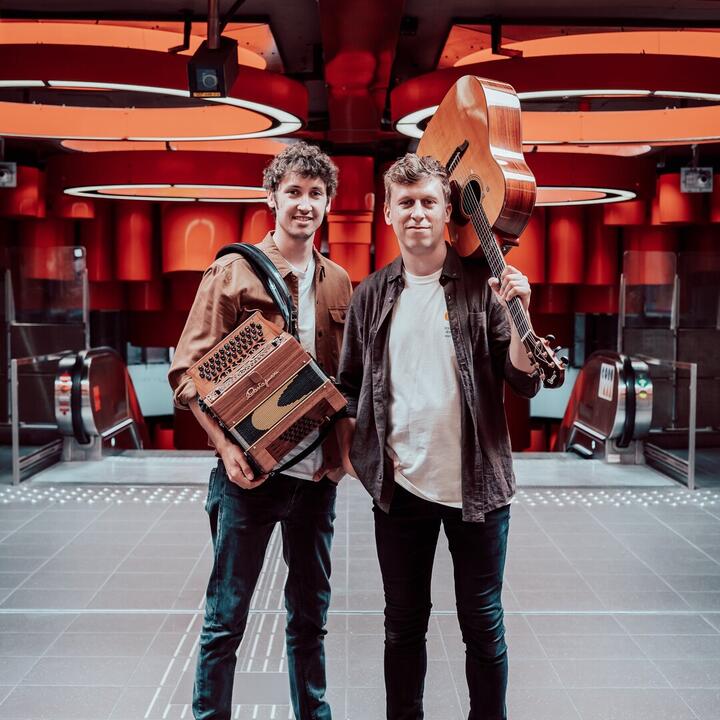 Thalas twee mensen met een accordeon en een gitaar in een fel verlichte ruimte met roltrappen.