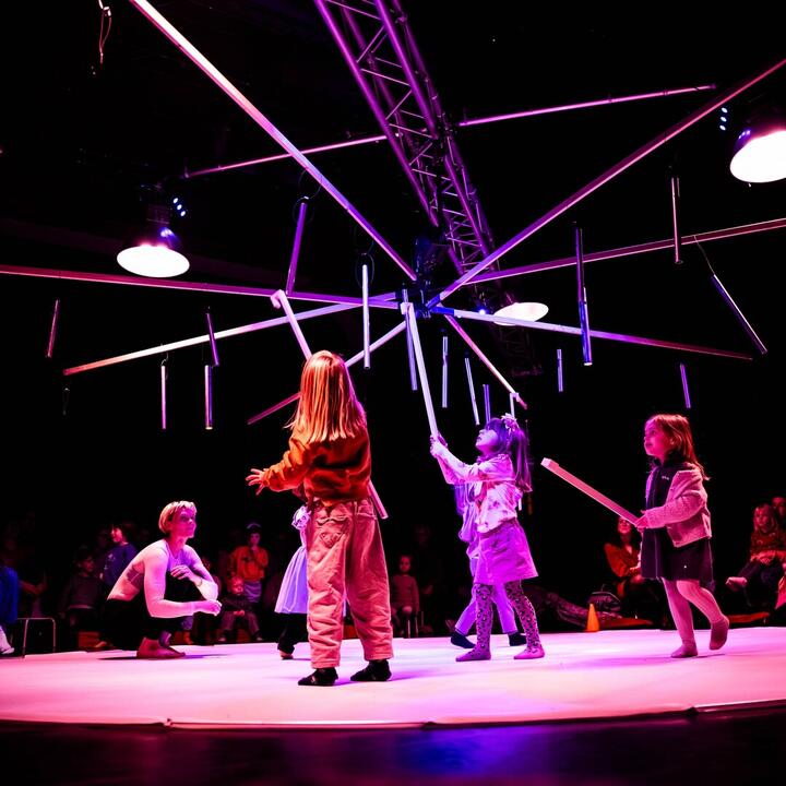Kinderen spelen op een verlicht podium onder een installatie van hangende staven en lampen, terwijl publiek rondom toekijkt. De scène baadt in roze en paarse theaterverlichting.