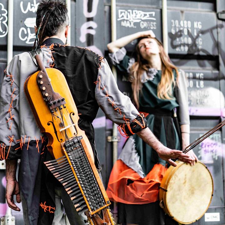 De afbeelding toont twee personen in kostuums met een nyckelharpa en een trommel, voor een container met graffiti.