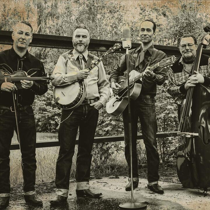Vier personen buiten, elk met een muziekinstrument: een viool, een banjo, een gitaar en een contrabas. Er staat een microfoonstandaard voor hen, wat suggereert dat ze optreden of zich voorbereiden op een muzikale uitvoering. De zwart-witkleuren geven de foto een vintage uitstraling.