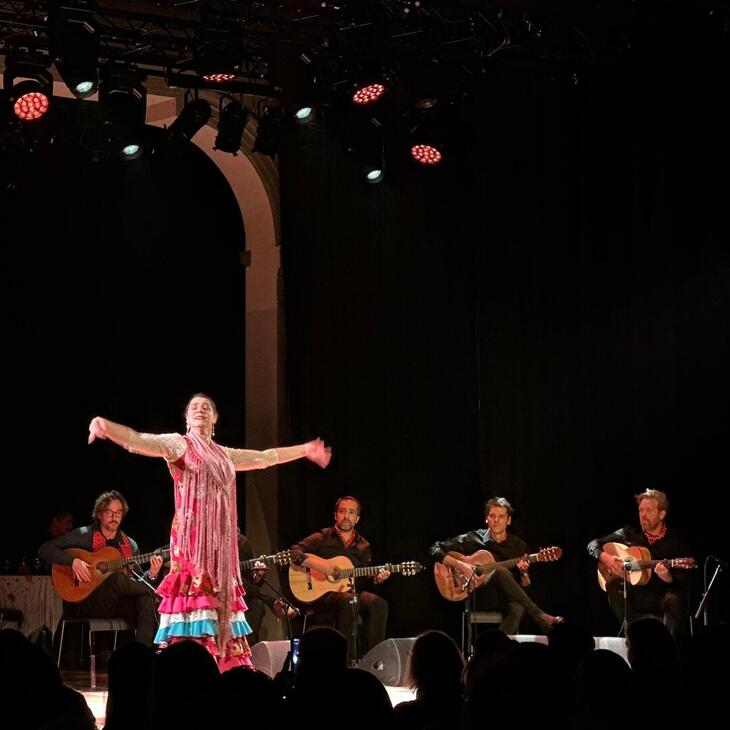 Een mevrouw staat vooraan. Achter haar zitten 5 mannen met een gitaar.