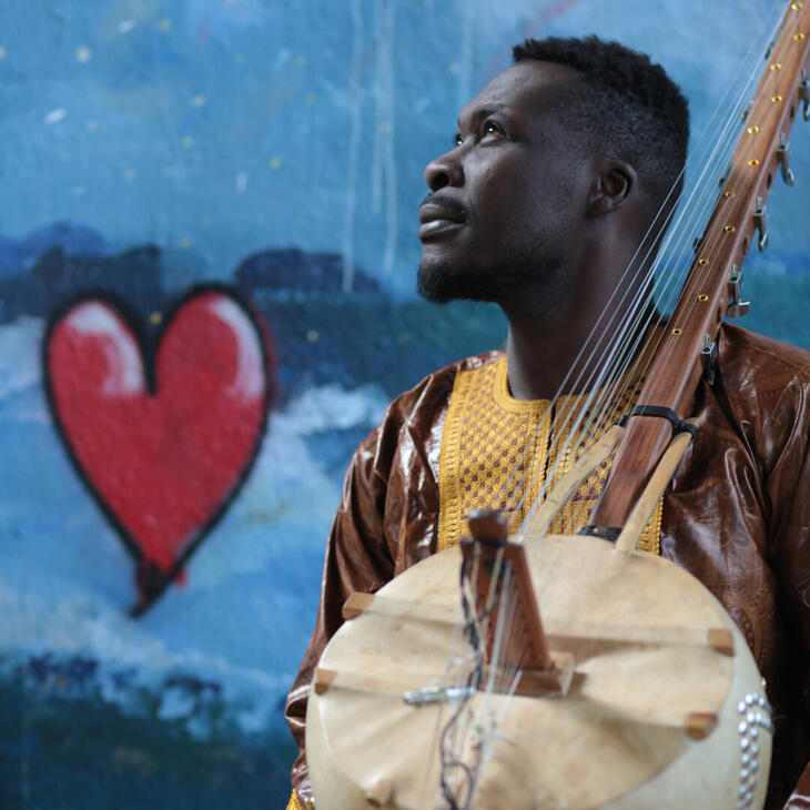 Aboubacar Traoré De afbeelding toont een persoon die een traditioneel snaarinstrument vasthoudt, met een achtergrond van een geschilderde muur met een groot rood hart en abstracte blauwe en witte patronen.