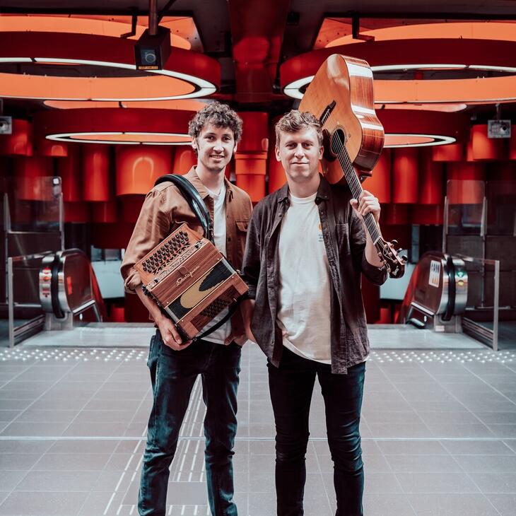 Thalas twee mensen met een accordeon en een gitaar in een fel verlichte ruimte met roltrappen.